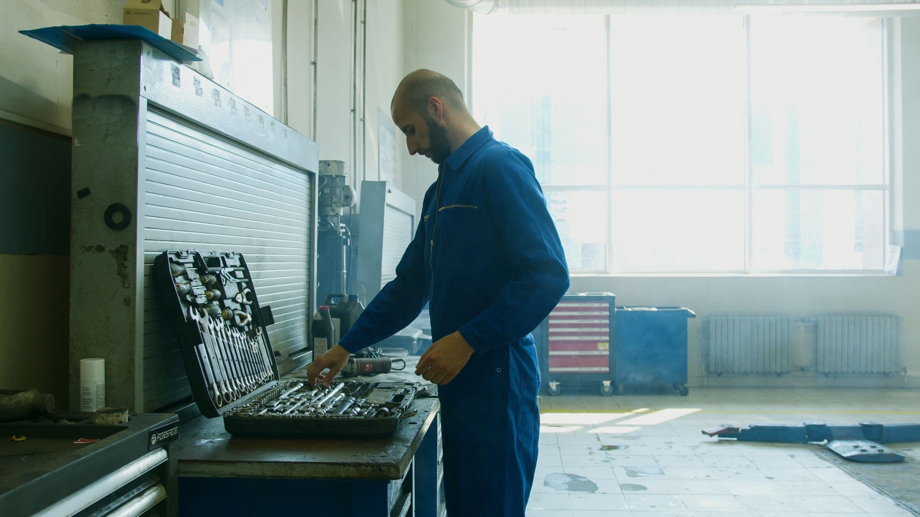 A mechanic in blue coveralls at a workshop bench with an open socket set and a red rolling tool cabinet behind