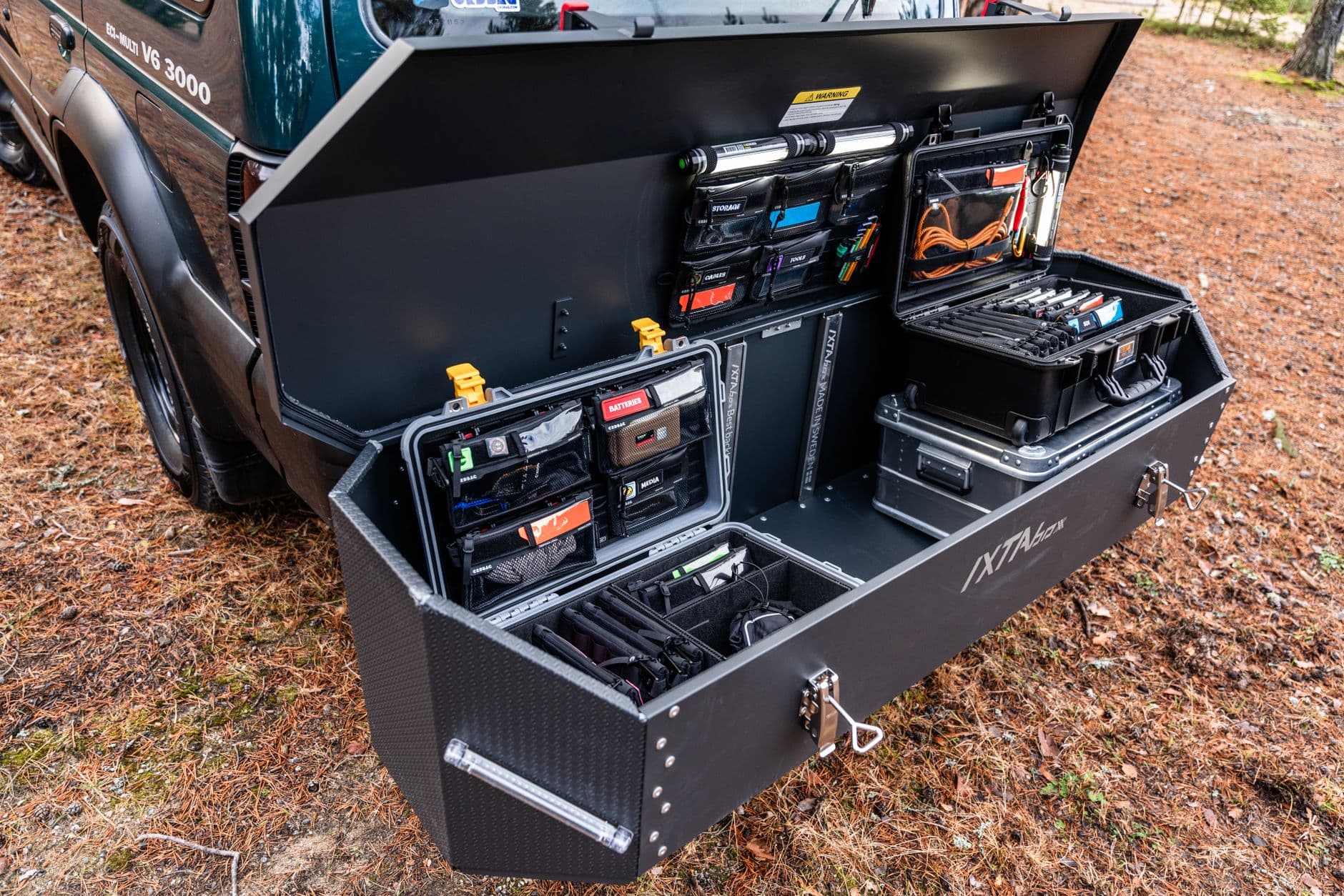 A vehicle-mounted storage drawer open at the tailgate, holding hard cases fitted with dividers and labeled organizer panels