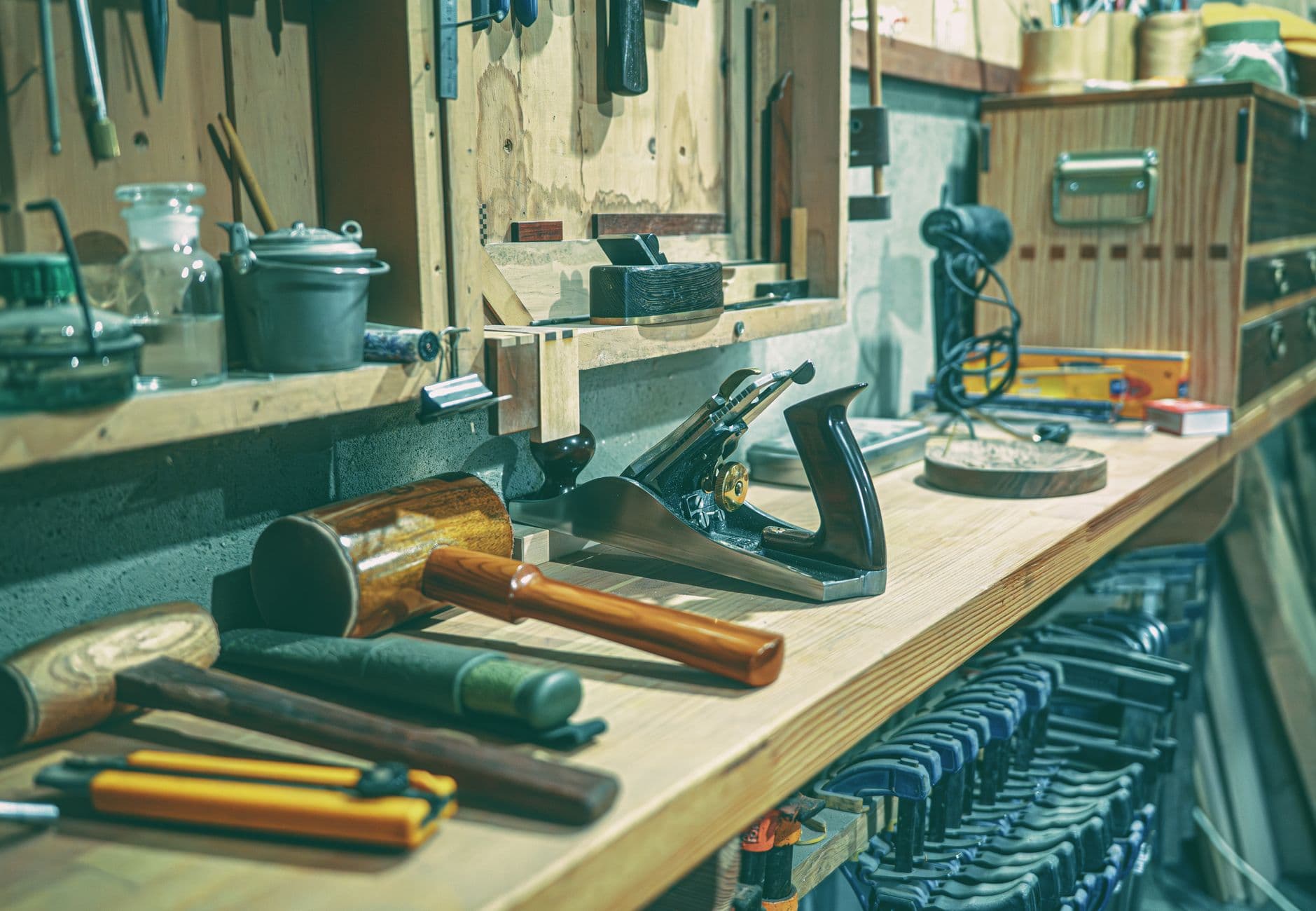 A wooden workbench top with hand planes, mallets and chisels laid out, a drawer chest behind it and clamps racked underneath