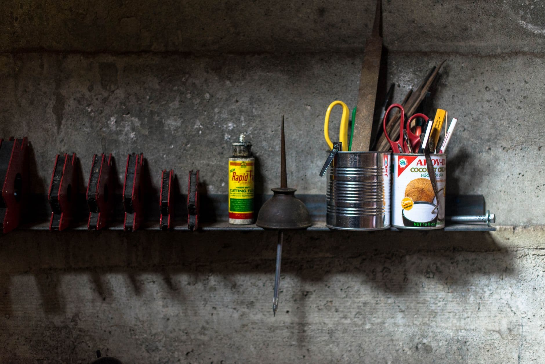 A narrow steel shelf on a workshop wall holding an oil can, cutting fluid and tins of small tools