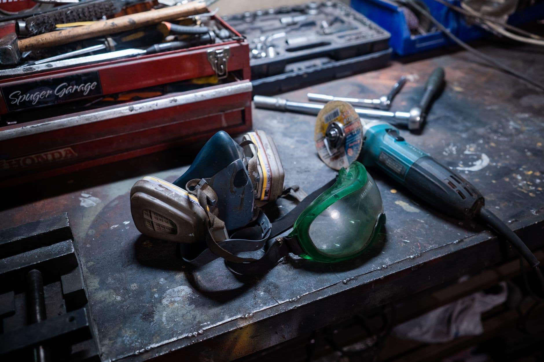 A bare metal workbench surface holding a red tool box, a socket set, an angle grinder and safety gear